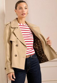 Beige doppelreihiger Trenchcoat mit glänzenden Knöpfen und Taschen, mit Leoprint gefüttert. Kombiniert mit einem rot-weiß gestreiften Hemd und dunklen Jeans.