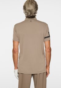 Mężczyzna stojący tyłem, ubrany w beżową koszulkę polo z czarnym logo na górnej części pleców i paskami na lewym rękawie, beżowe spodnie do kompletu oraz białą rękawiczkę.