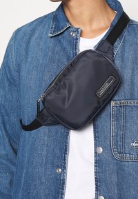Mężczyzna w niebieskiej dżinsowej kurtce i białej koszuli, z czarną torbą na ramię Calvin Klein przypasaną na klatce piersiowej.
