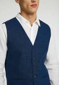 Temno modra lanena majica z diskretno teksturo, z V-izrezom in šestimi črnimi gumbi; nosi se čez belo srajco z ovratnikom.