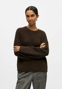 Pull en tricot marron à coupe décontractée, avec un col et des poignets côtelés, assorti à un pantalon à carreaux gris avec une silhouette ample.