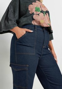 Farkku cargo-housut korkealla vyötäröllä, syvän sininen väri, etu- ja sivutaskut, sekä vastakkaiset oranssit ompelu- ja yksityiskohtateokset.