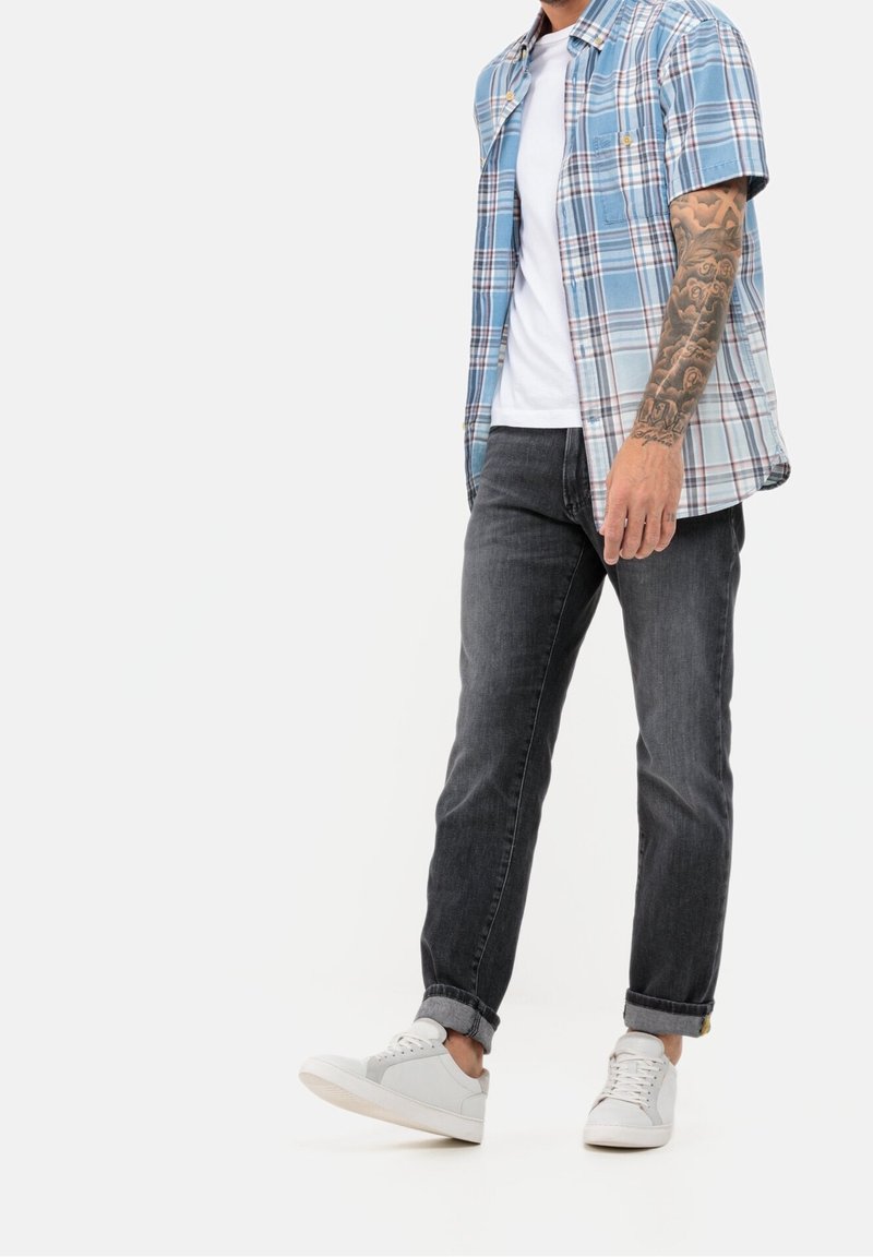 Homme portant un jean noir retroussé, des baskets blanches, un t-shirt blanc et une chemise à manches courtes bleue à carreaux ouverte, avec un bras droit tatoué visible.
