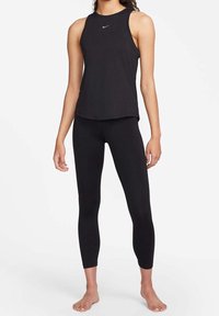 Žena stojící bosá, oblečená v černém bezramínkovém topu Nike a černých zkrácených legínách na bílém pozadí.