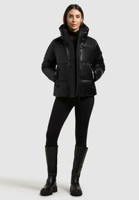 Sort pufferjakke med høj krave, quiltet design, blankt materiale, frontlommer og lynlåslukning, båret over sorte leggings og knælange støvler.