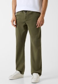 Olivgrüne Baumwollhose mit lockerem Sitz, elastischem Bund und verstellbarem Kordelzug. Kombiniert mit weißen Turnschuhen. Glatte Textur.