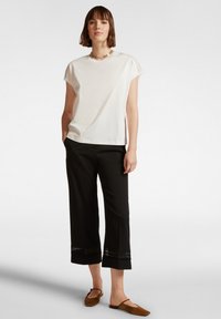 T-shirt bianco in cotone con dettagli in pizzo sulle spalle, abbinato a pantaloni neri a 3/4 con bordo in pizzo all'orlo, e scarpe slip-on marroni.