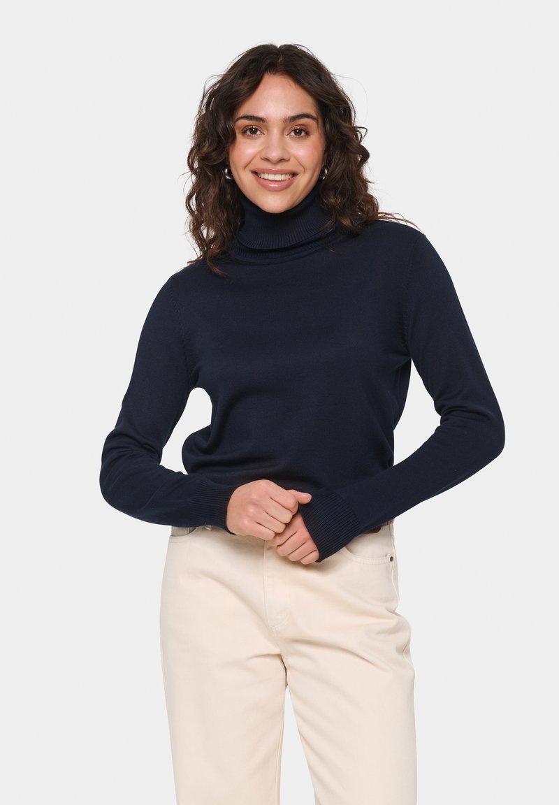 Navy blue turtleneck sweater with ribbed texture and long sleeves, paired with light-colored pants, hands placed at the waist.