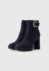 Une paire de bottes de cheville en suède noir avec des semelles compensées et un talon bloc, ornées d'une sangle à boucle dorée sur fond blanc.