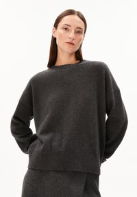 Frau mit glatten braunen Haaren trägt einen dunkelgrauen, übergroßen Pullover und einen passenden Rock und posiert mit den Händen auf dem Rücken vor einem weißen Hintergrund.