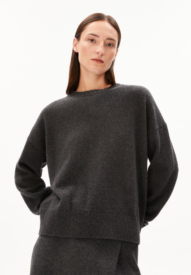 Frau mit glatten braunen Haaren trägt einen dunkelgrauen, übergroßen Pullover und einen passenden Rock und posiert mit den Händen auf dem Rücken vor einem weißen Hintergrund.