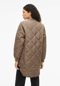 Ruda dygsniuota striukė su rombo rašto raštu, ribinėmis rankogaliais ir apvaliomis apačiomis. Nugara matoma sklandži tekstūra ir laisvas siluetas.