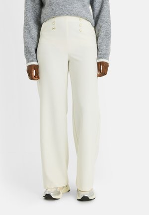 Persona vistiendo pantalones crema de pierna ancha de talle alto con cuatro botones, suéter gris de punto y zapatillas deportivas blancas plateadas sobre fondo blanco.