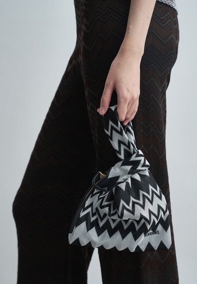 Fekete-fehér cikkcakk mintájú táska, amelyet egy kéz tart, csomózott füllel, sötét cikkcakk mintájú nadrágot viselve, sima háttér előtt.