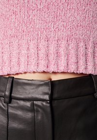 Gros plan sur un ventre portant un pull court texturé rose et un pantalon en cuir noir taille haute avec passants de ceinture.