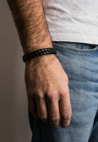 Melnas austu rokas josla, kas izgatavota no ādas, ar strukturētu dizainu un gludu tekstūru, uz plaukstas ar redzamām matu šķipsnām.