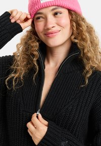 Pull en tricot noir avec un col haut et une fermeture éclair, présentant une texture côtelée et des manches larges. Le modèle porte un bonnet rose.