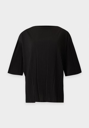 Femme portant un haut noir et un pantalon beige à jambes larges tenant un sac à main taupe, debout devant un fond clair uni.