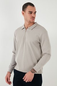 Vīrietis ar īsiem matiem, tērpies gaiši pelēkā garroku polo krekliņā ar īsu rāvējslēdzēju, skatās uz labo pusi, rāda rokas tetovējumus.