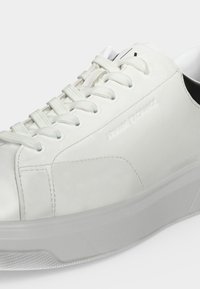 Gros plan d'une basket en cuir blanc avec des lacets blancs, des coutures détaillées, une semelle épaisse et un accent noir sur le talon.