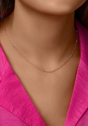 Collar de cadena en oro rosa con pequeños acentos redondos, llevado sobre una blusa rosa vibrante. Presenta un diseño delicado y minimalista.