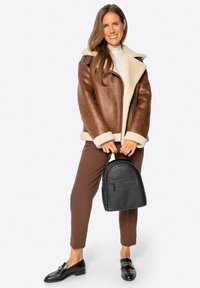 Braune Kunstlederjacke mit Lammfellfutter, braune Hose, schwarzer Nieten-Rucksack und schwarze Mokassins. Das Model steht lächelnd.