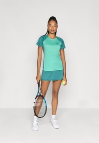 Tealne sportlik riietus lühikeste varrukatega särgi ja seeliku kombinatsioonis. Omab sujuvat tekstuuri ja kontrastseid varrukaid. Käes on tennisereket ja pall.
