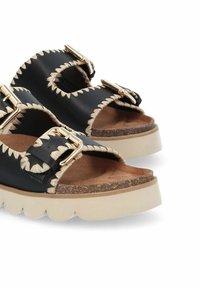 Sandales noires en cuir avec deux lanières, boucles dorées et accents tissés beiges. Semelle plate-forme blanche épaisse avec une semelle intérieure texturée beige.