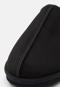 Papuc negru cu textură moale de suede, formă rotunjită și cusături contrastante pe margine, având o talpă căptușită.