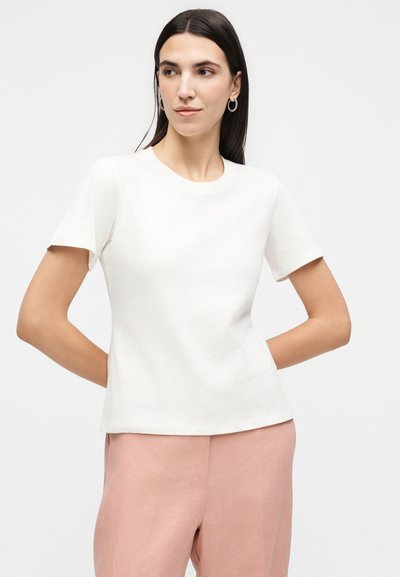 Γυναίκα με σκούρα μαλλιά που φορά ασημένια σκουλαρίκια στεφάνια, μια απλή λευκή κοντομάνικη μπλούζα και ανοιχτό ροζ παντελόνια, στέκεται με τα χέρια πίσω από την πλάτη.