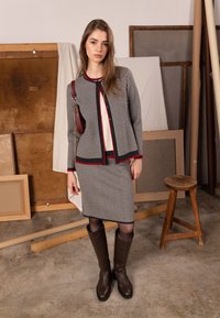 Šedé a bílé kostkované sako s červeným lemem, doplněné o odpovídající sukni. Průsvitné černé punčocháče a vysoké hnědé kožené boty.