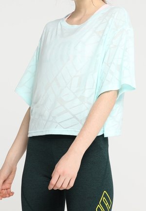 Kvinde iført en løs, lyseblå, mønstret kortærmet top og mørke tætsiddende leggings med et gult logo på låret.