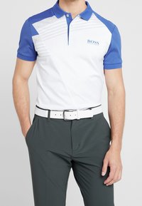 Kortärmad polo i vitt och blått, med diagonala designelement, texturerad tyg och en logotyp på vänstra bröstet. Matchas med mörka byxor och ett vitt bälte.