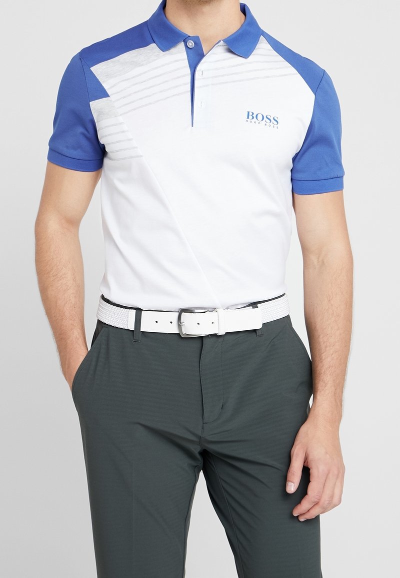 Kortärmad polo i vitt och blått, med diagonala designelement, texturerad tyg och en logotyp på vänstra bröstet. Matchas med mörka byxor och ett vitt bälte.