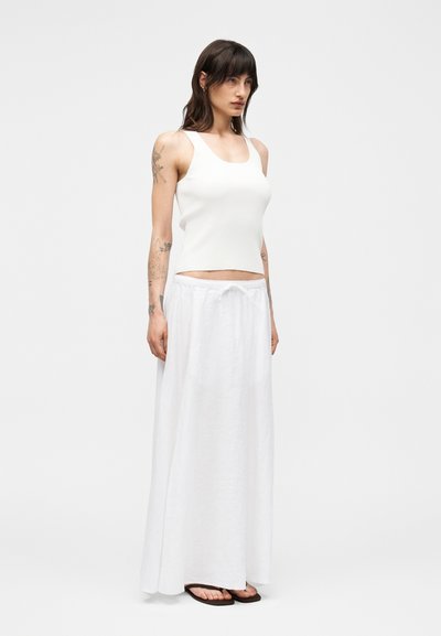 Γυναίκα με τατουάζ που φοράει λευκό αμάνικο μπλουζάκι, μακριά λευκή φούστα με κορδόνι στη μέση και καφέ σαγιονάρες, στέκεται μπροστά σε απλό φόντο.