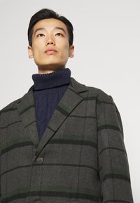 Ull-blandet frakk med grå og grønn rutete mønster, enkeltknappede design, hakkete slag og knappestenging, båret over en mørkeblå turtleneck.
