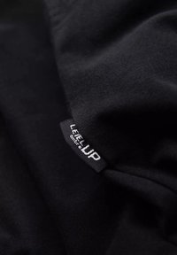 Černá látka s jemnou texturou a obdélníkovou nálepkou s nápisem "LEVEL UP" v bílé. Materiál se zdá být hladký a mírně elastický.