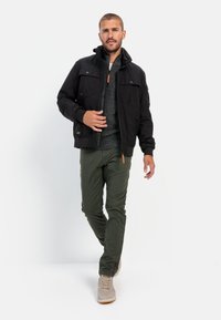 Black zip-up jacket with a high collar, two chest pockets, paired with a dark knitted sweater and olive green pants, finished with light sneakers.