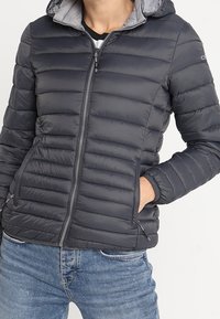 Chaqueta acolchada gris oscuro con cremallera frontal, capucha y puños acanalados. Presenta compartimentos horizontales, bolsillos laterales y un forro interior liso.