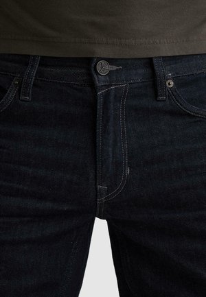 Nahaufnahme der Taille einer dunkelblauen Jeans mit sichtbarem Knopf, Reißverschluss, Gürtelschlaufen und dem oberen Teil eines olivgrünen Hemdes.