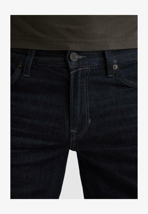 Nahaufnahme der Taille einer dunkelblauen Jeans mit sichtbarem Knopf, Reißverschluss, Gürtelschlaufen und dem oberen Teil eines olivgrünen Hemdes.