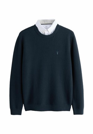 Mørkeblå sweater med en struktureret vævning, rund hals og lange ærmer, prydet med et lille broderet logo på brystet og lagdelt over en hvid skjorte med krave.