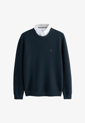 Pull en bleu marine foncé avec un tissage texturé, col rond et manches longues, arborant un petit logo brodé sur la poitrine et superposé à une chemise blanche col.