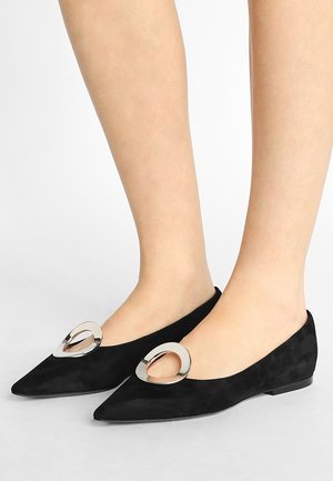 Zapatos planos de ante negro con punta puntiaguda y una gran hebilla plateada en forma de lágrima, usados en pies descalzos sobre fondo blanco.