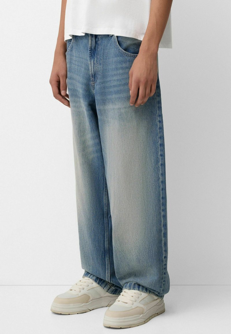 Helle blaue baggy Jeans mit ausgeblichenem Washed-Effekt, Fünf-Taschen-Design und sichtbaren Nähten, kombiniert mit hellen Sneakers.
