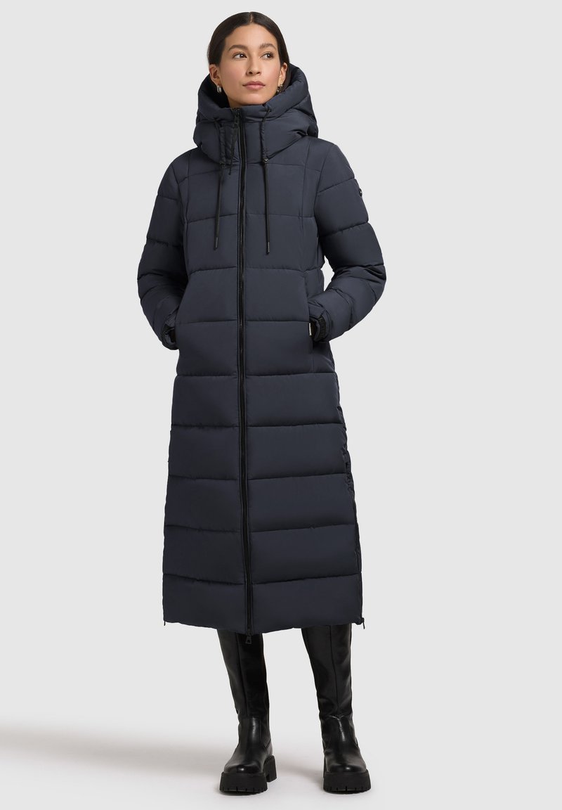 Cappotto imbottito lungo di un blu navy scuro con cappuccio, dotato di zip frontale, motivo a trapunta e tasche laterali, abbinato a stivali neri alti fino al ginocchio.