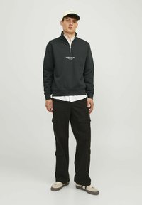 Sudadera negra con cremallera y cuello alto, con texto de logo blanco. Combinada con pantalones cargo oscuros y zapatillas beige. Diseño simple en general.