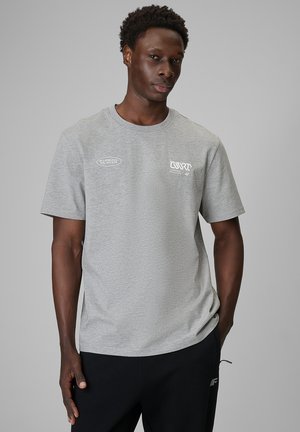 Mężczyzna stoi z rękami w kieszeniach czarnych spodni, nosi jasnoszary T-shirt z białymi logo na piersi, neutralne szare tło.