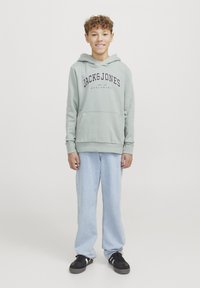 Hoodie verde chiaro con stampa "JACK&JONES", tasca a marsupio, abbinato a jeans larghi azzurri e sneakers nere.