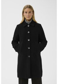 Schwarzer Wollmantel mit kragenförmigem Ausschnitt, großen Knöpfen, zwei seitlichen Taschen und taillierter Passform. Über einem grauen Rollkragenpullover getragen.